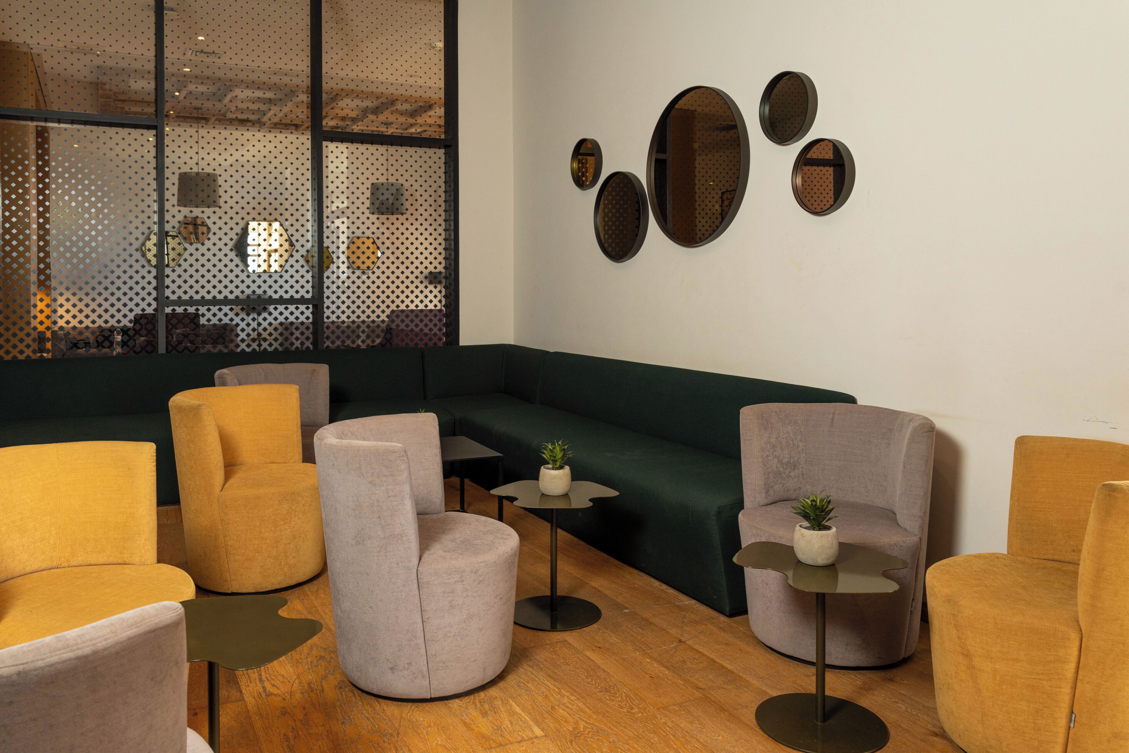 A modern lounge area with sofas, armchairs, and small tables, featuring a large window and decorative wall mirrors.