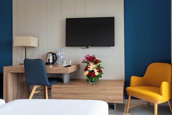 A hotel room with a bed, a desk with a chair, a TV, a vase with flowers, and a yellow armchair.