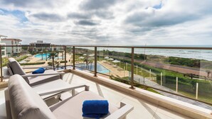 Un balcón con vista a una piscina, sillones de descanso y una playa lejana.