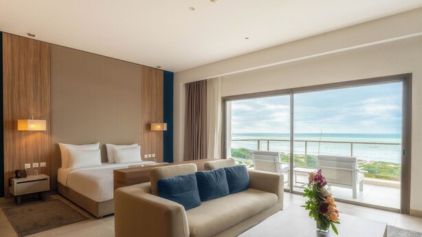 Una habitación de hotel moderna con cama, sofá, mesa de centro y un gran ventanal con vista al océano.