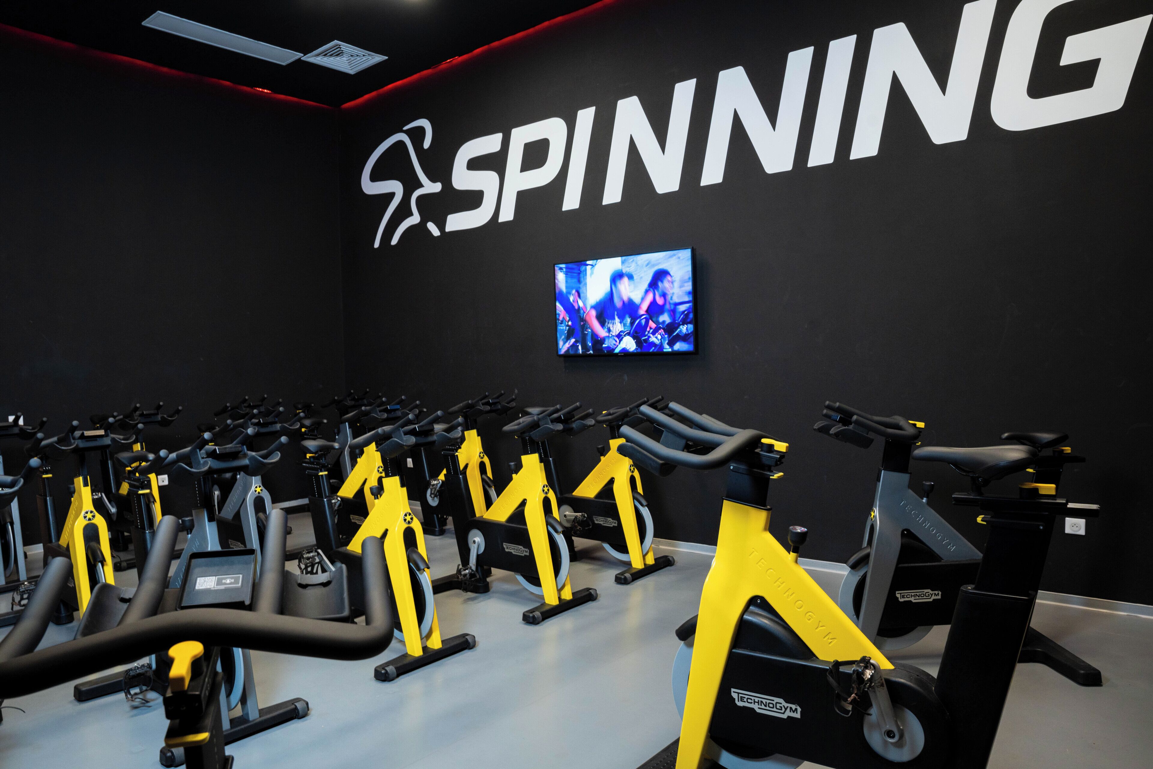 A gym with multiple stationary bikes, a large 'SPINNING' sign, and a TV screen displaying an image.