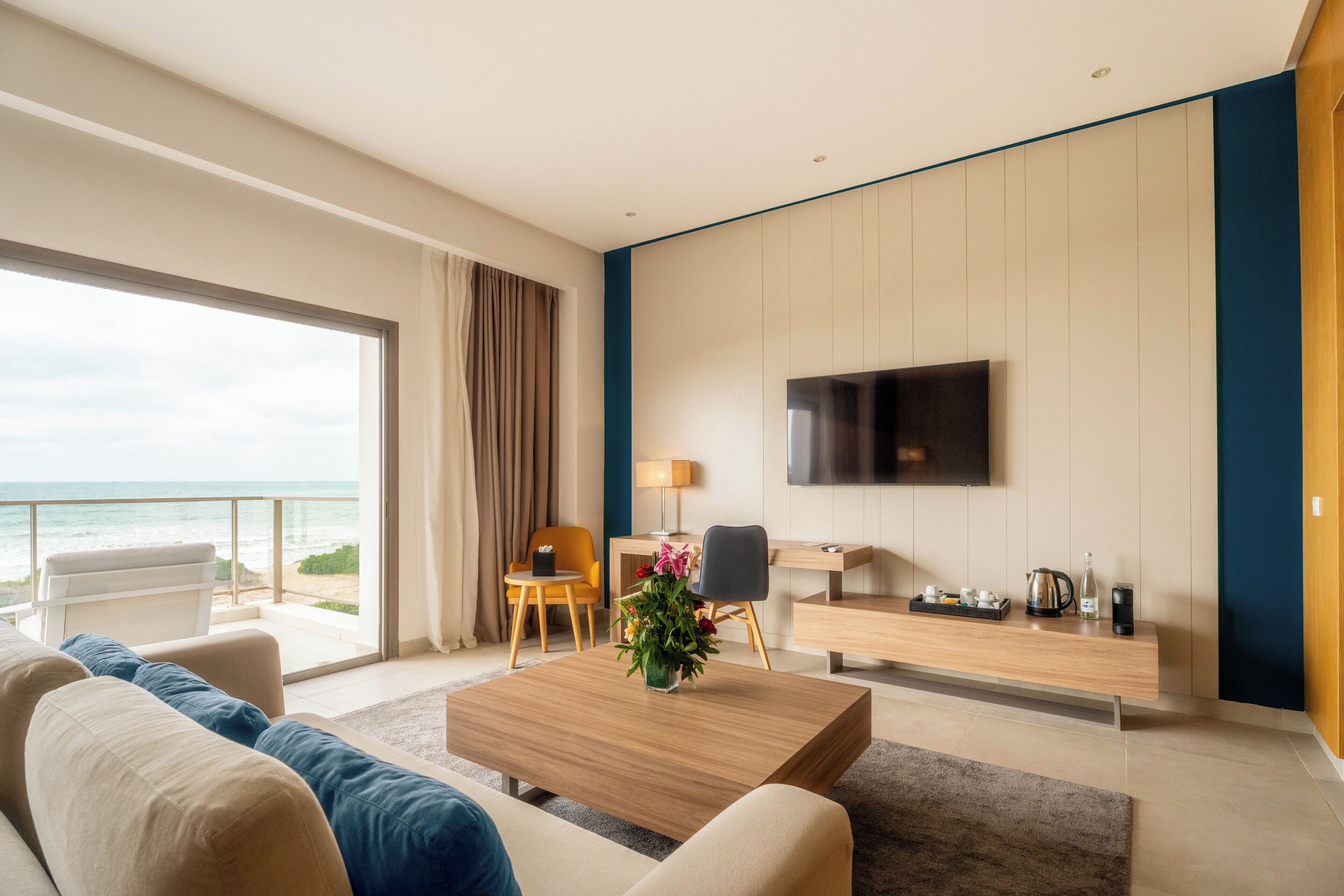 A modern hotel room with a sofa, coffee table, TV, desk, and a view of the ocean.