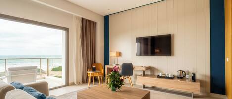 A modern hotel room with a sofa, coffee table, TV, desk, and a view of the ocean.