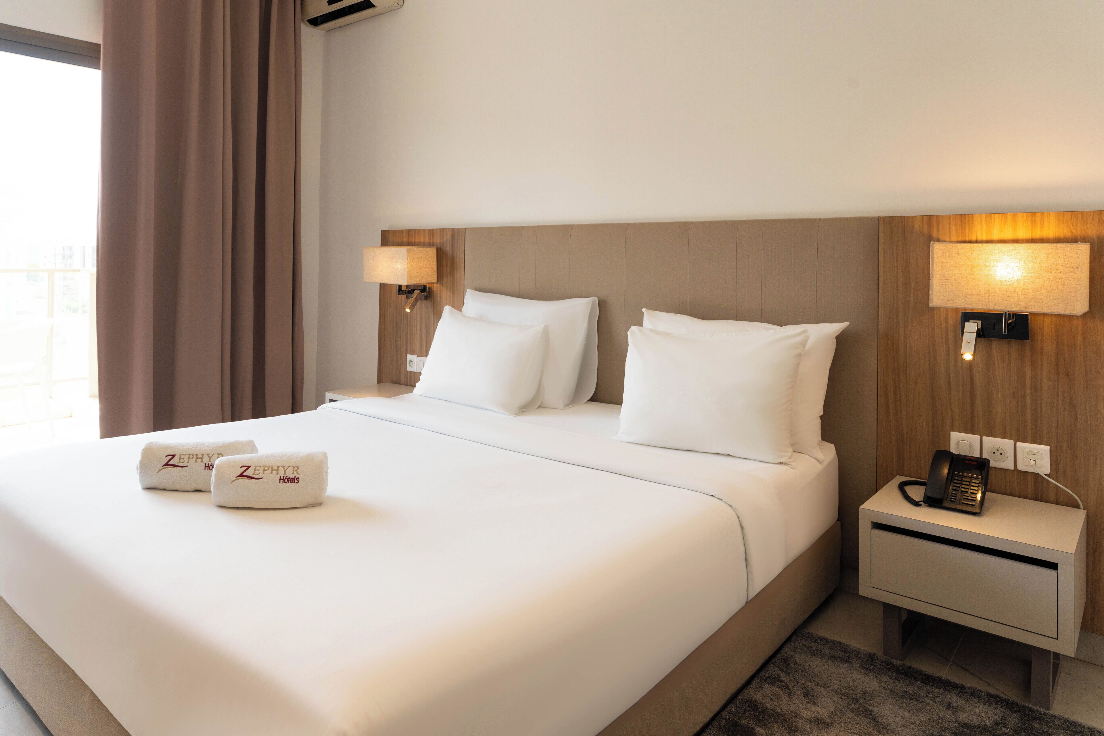 A hotel room with a large bed, two bedside lamps, a nightstand with a phone, and a window with curtains.