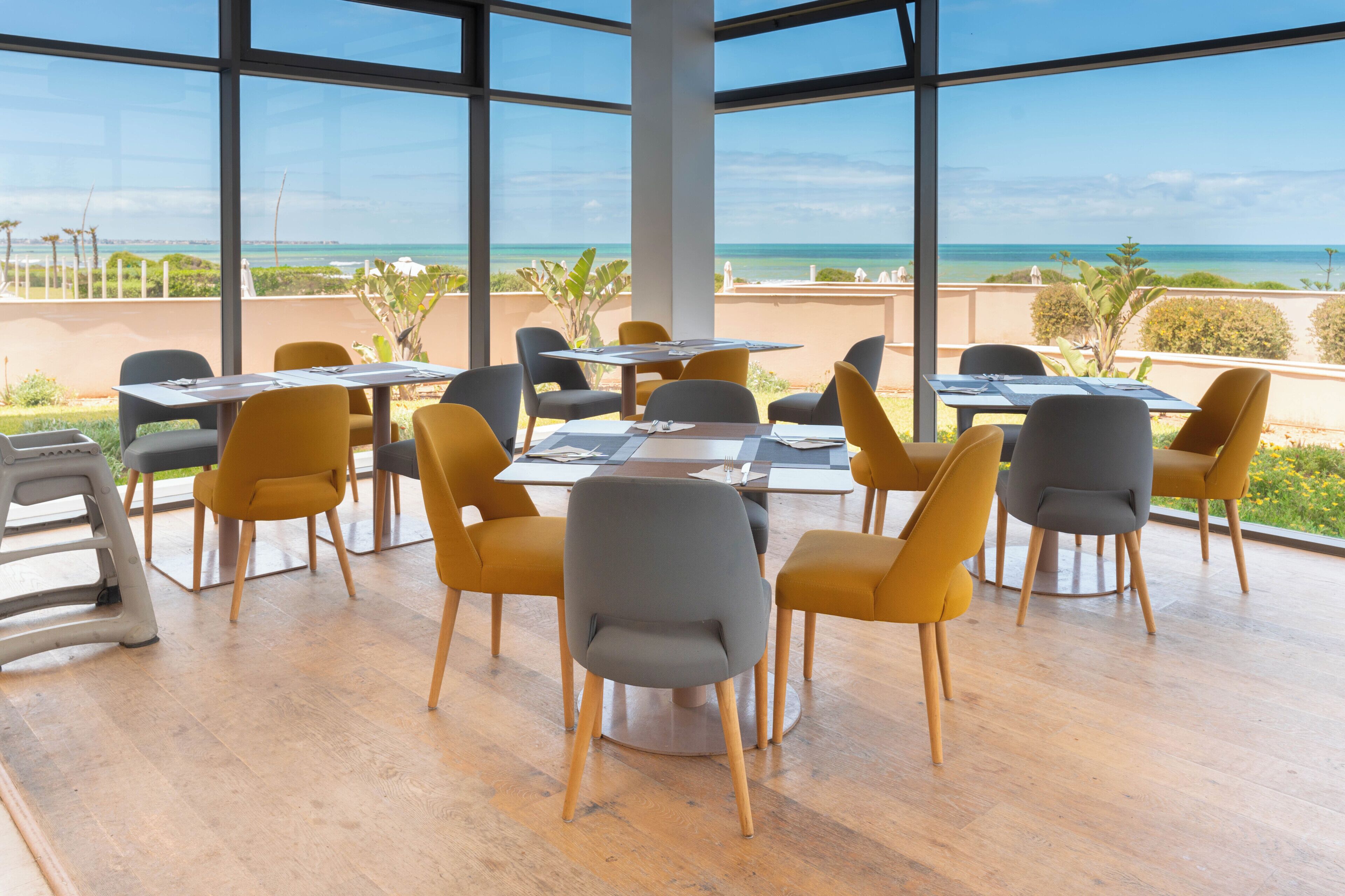 Un restaurante con mesas y sillas dispuestas cerca de amplios ventanales con vista a una playa.