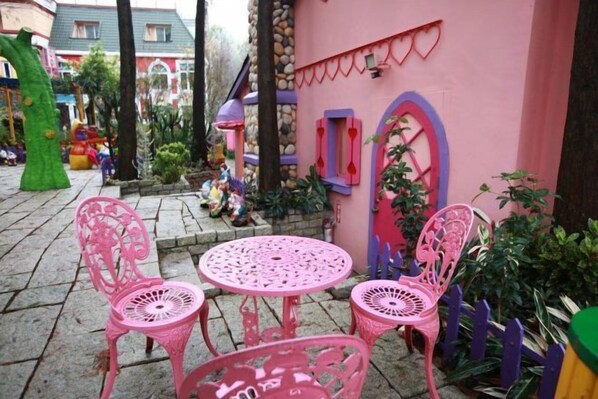 Un espace extérieur aménagé en salon, avec des chaises et une table, le tout dans des tons roses, et entouré de plantes et d’une clôture décorative.