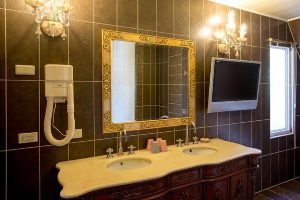 Une salle de bain avec deux lavabos, un grand miroir orné d’un cadre décoratif, un lustre et un téléviseur fixé au mur.
