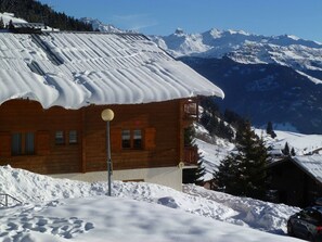 A wooden cabin with a snow-covered roof, a balcony, and a view of snow-covered mountains in the background.