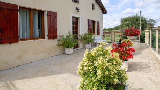 Una casa de dos pisos con fachada color beige, contraventanas de madera y un pequeño porche con plantas en macetas y un banco.