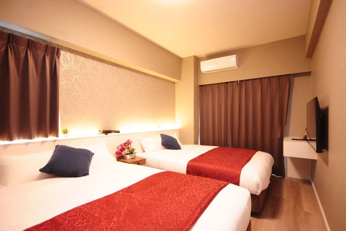 Habitación de hotel con dos camas, cada una con una manta roja, una mesita con lámpara, una planta y un televisor sobre un soporte.
