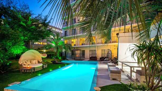 Una piscina con mobiliario exterior y vegetación, un edificio de varios pisos con balcones, y un patio bien iluminado.