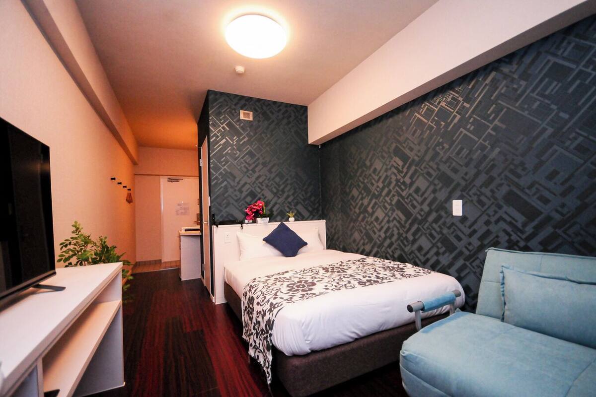 Una habitación de hotel con cama, escritorio, televisión, silla y un papel tapiz estampado.