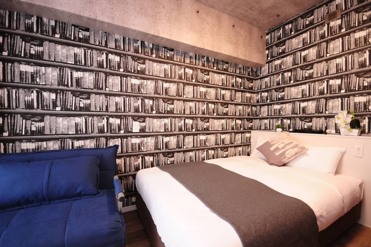 Un dormitorio con una gran estantería llena de libros, una cama con ropa de cama blanca y marrón, y un sofá azul.