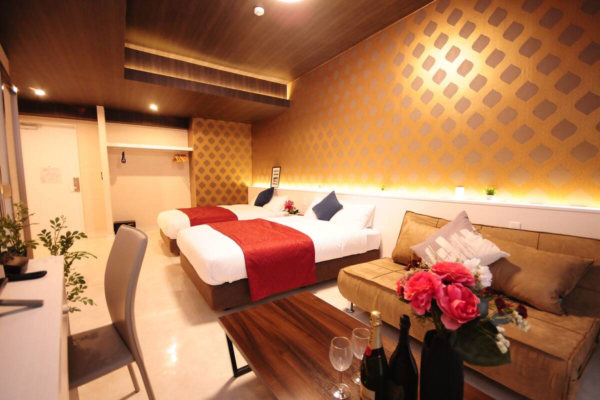 Habitación de hotel con dos camas, un sofá, una mesa de madera y un arreglo floral, además de una planta pequeña.