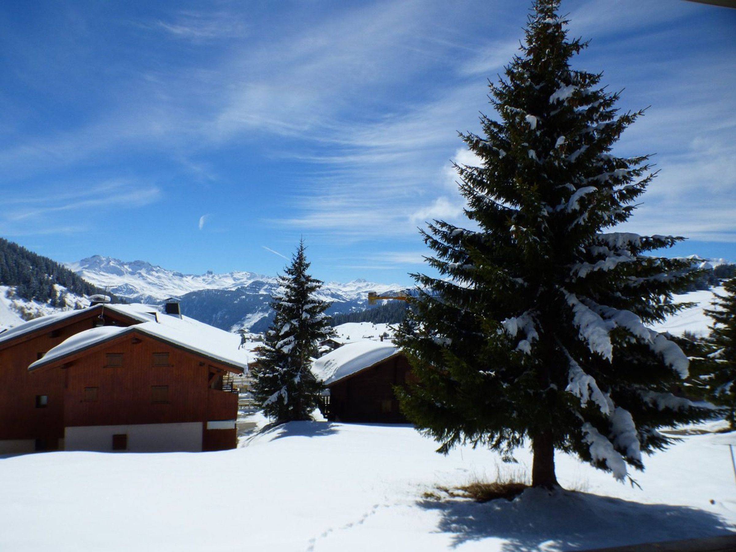 A wooden cabin with a snow-covered roof, surrounded by snow-covered ground and trees, with a mountain range in the background.