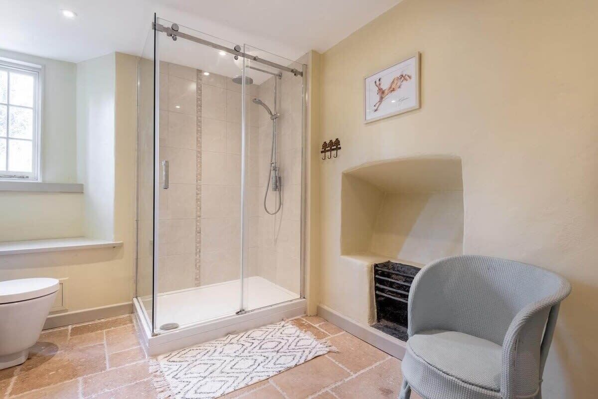 A bathroom with a glass-enclosed shower, a white toilet, a patterned rug, and a chair.