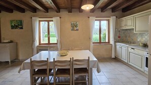 A dining room with a wooden ceiling, two windows with curtains, a table with chairs, and a kitchen area with cabinets and a sink. - Charming Perigord house in Salignac-Eyvigues, pets allowed (Salignac-Eyvigues)