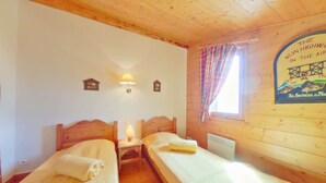 A room with two wooden beds, a small table with a lamp, a radiator, and a window with curtains. - 4-Room Mezzanine Apt Les Saisies 10 guests (Villard-sur-Doron)