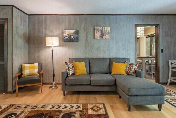 A living room with a grey sectional sofa, a wooden chair, a floor lamp, and two wall-mounted pictures.