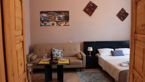 Una habitación de hotel con sofá, una mesita con una copa de vino y un plato, cama con almohadas y dos adornos en la pared.