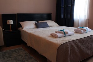 Een keurig opgemaakt bed met een beige dekbedovertrek en kussens, een nachtkastje met een lamp, en een raam met gordijnen.