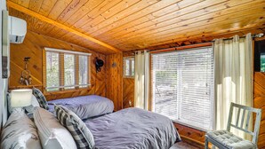 A wooden cabin room with a bed, two windows, a chair, and a wall-mounted air conditioner. - Near Lake + Trails: Rustic Newland Cabin w/ Deck! (Newland)