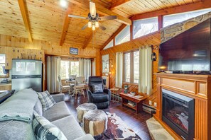 Interior acogedor de una cabaña con techo de madera, chimenea, sofá, zona de comedor y cocina con refrigerador.
