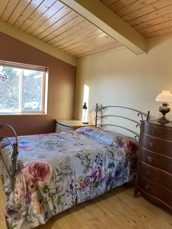 A bedroom with a wooden ceiling, a bed with floral bedding, a dresser, and a window with a view of trees. - Homer, Alaska 4BR House-great views, dogs welcome, close to town (Homer)