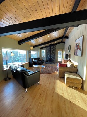 A spacious living room with wooden floors, a stone fireplace, and a large window offering a view of the outdoors. - Homer, Alaska 4BR House-great views, dogs welcome, close to town (Homer)