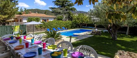 A poolside dining area with a table set for a meal, surrounded by chairs and a view of a swimming pool and mountains in the background.