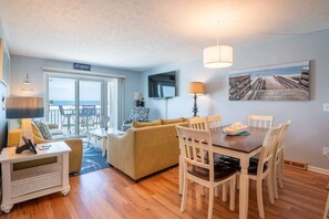 A cozy living room with a sofa, dining table, and chairs. A TV is mounted on the wall, and there is a window with a view of the beach. - Ocean Dunes 2005 Wait N Sea (Kure Beach)