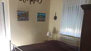 A bedroom with a bed, a chandelier, two paintings, a radiator, and a window with curtains.