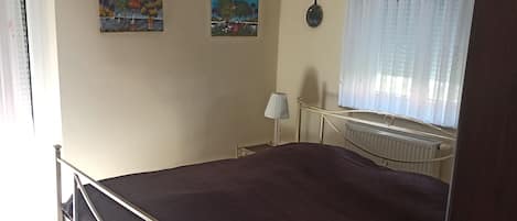 A bedroom with a bed, a chandelier, two paintings, a radiator, and a window with curtains.