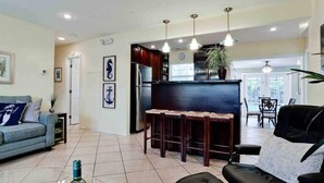 A modern living room with a sofa, a coffee table, and a bar area with stools. There is a refrigerator, a dining table with chairs, and a large window providing natural light.
