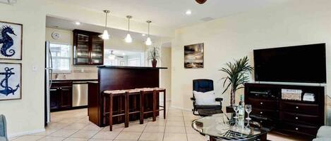 A modern living room with a kitchen, a bar area, a television, and a ceiling fan.