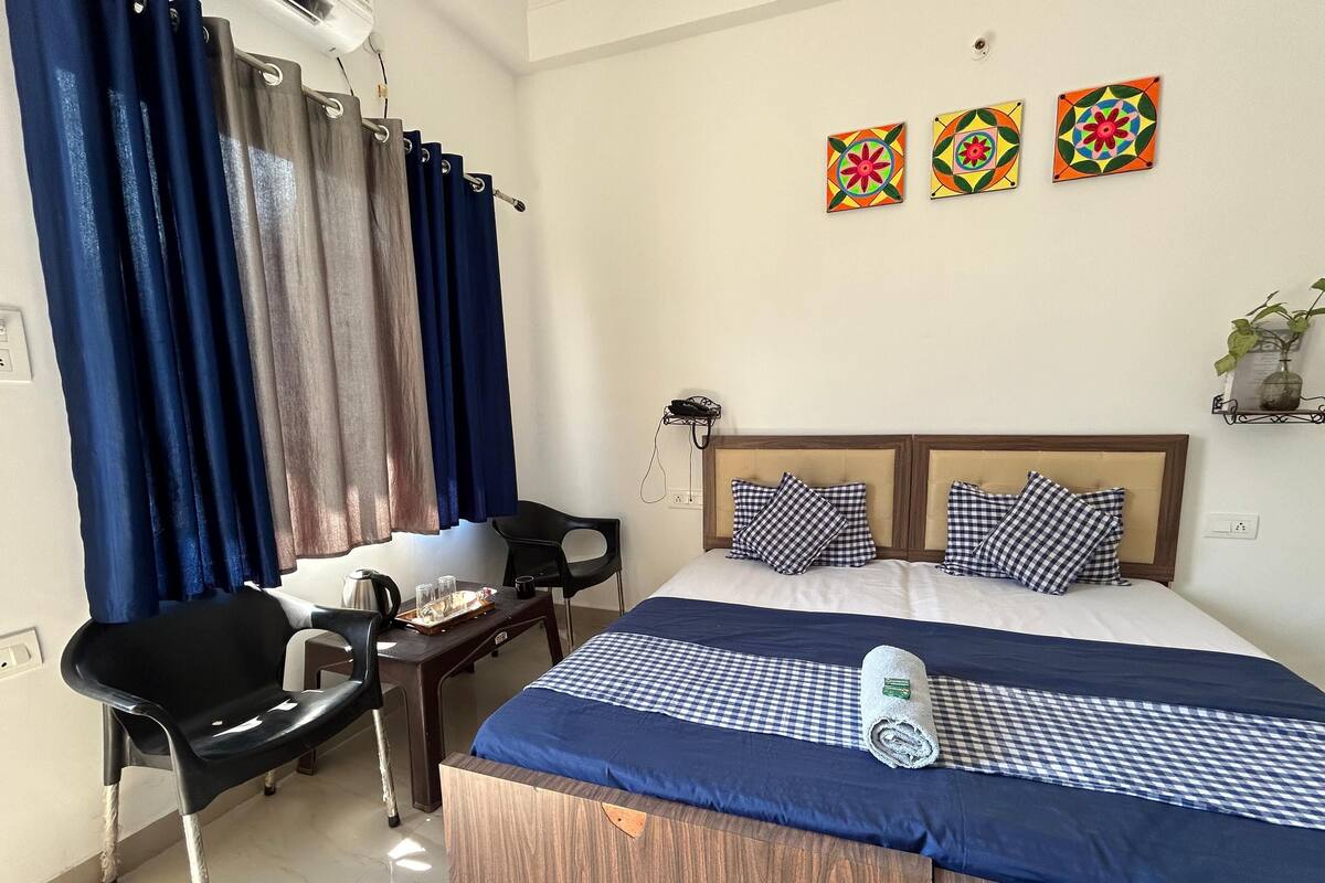 A hotel room with a bed, a desk with a chair, a coffee maker, and a small table. There are three decorative wall pieces above the bed.