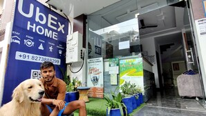 A hotel entrance with a large blue banner advertising 'UberX Home' services, a contact number, and amenities like food, tours, and pet-friendly options. - UbEx Home Rishikesh - Hostel (Narendranagar)