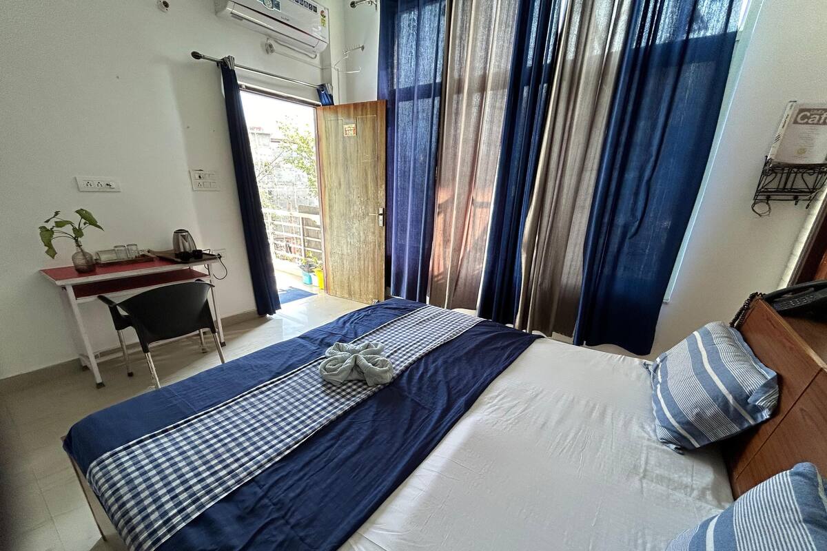 A hotel room with a bed, a desk, and a chair. There is a window with curtains, and an air conditioning unit on the wall.