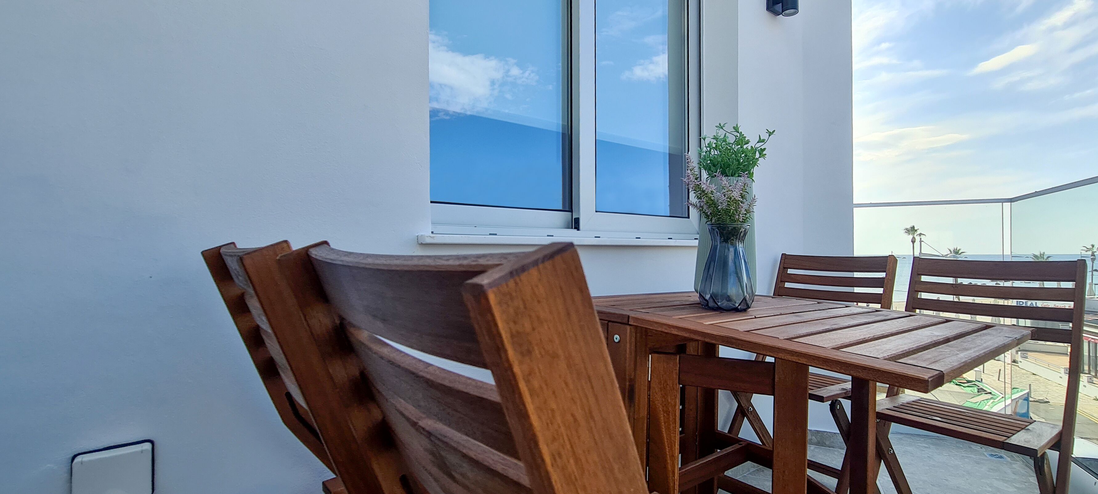 Une table et des chaises en bois sont placées devant un mur blanc. Une fenêtre et un vase de fleurs ornent la table.