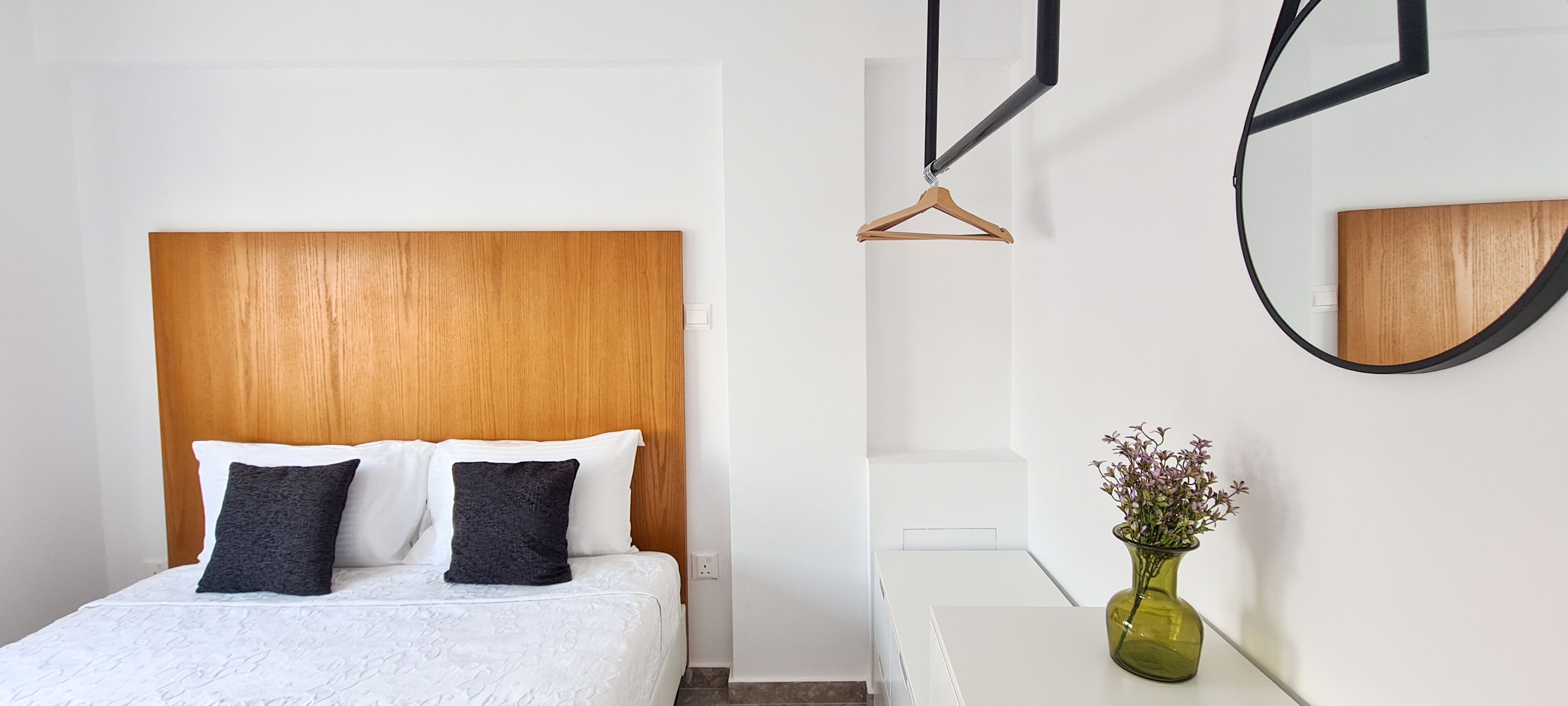 Habitación de hotel con cabecera de madera, cama blanca con dos almohadas, mesita de noche con un florero y un espejo en la pared.