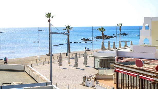 A beachside view with palm trees, a clear blue sea, and a building with a red awning. - Phaedrus Living: Seaside Luxury Flat Lighthouse 69 (Paphos)