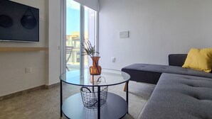 A modern living room with a glass table, a grey sofa with a yellow pillow, and a large TV mounted on the wall. - Phaedrus Living: Seaside Luxury Flat Lighthouse 69 (Paphos)