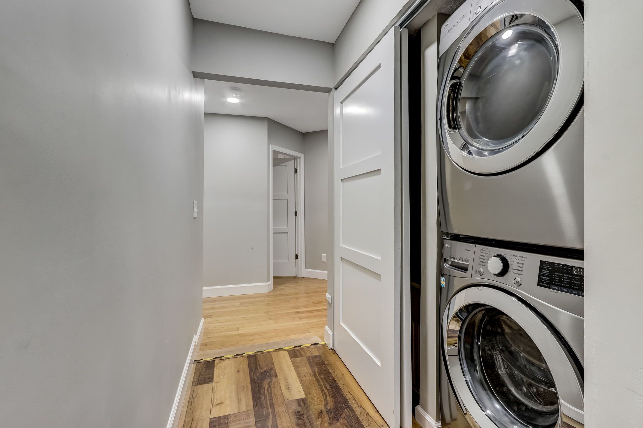 A laundry room with two stacked front-loading washing machines, a wooden floor, and a white door leading to another room.
