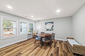 Un coin repas comprenant une table ronde en bois, six chaises et une grande fenêtre avec des stores.