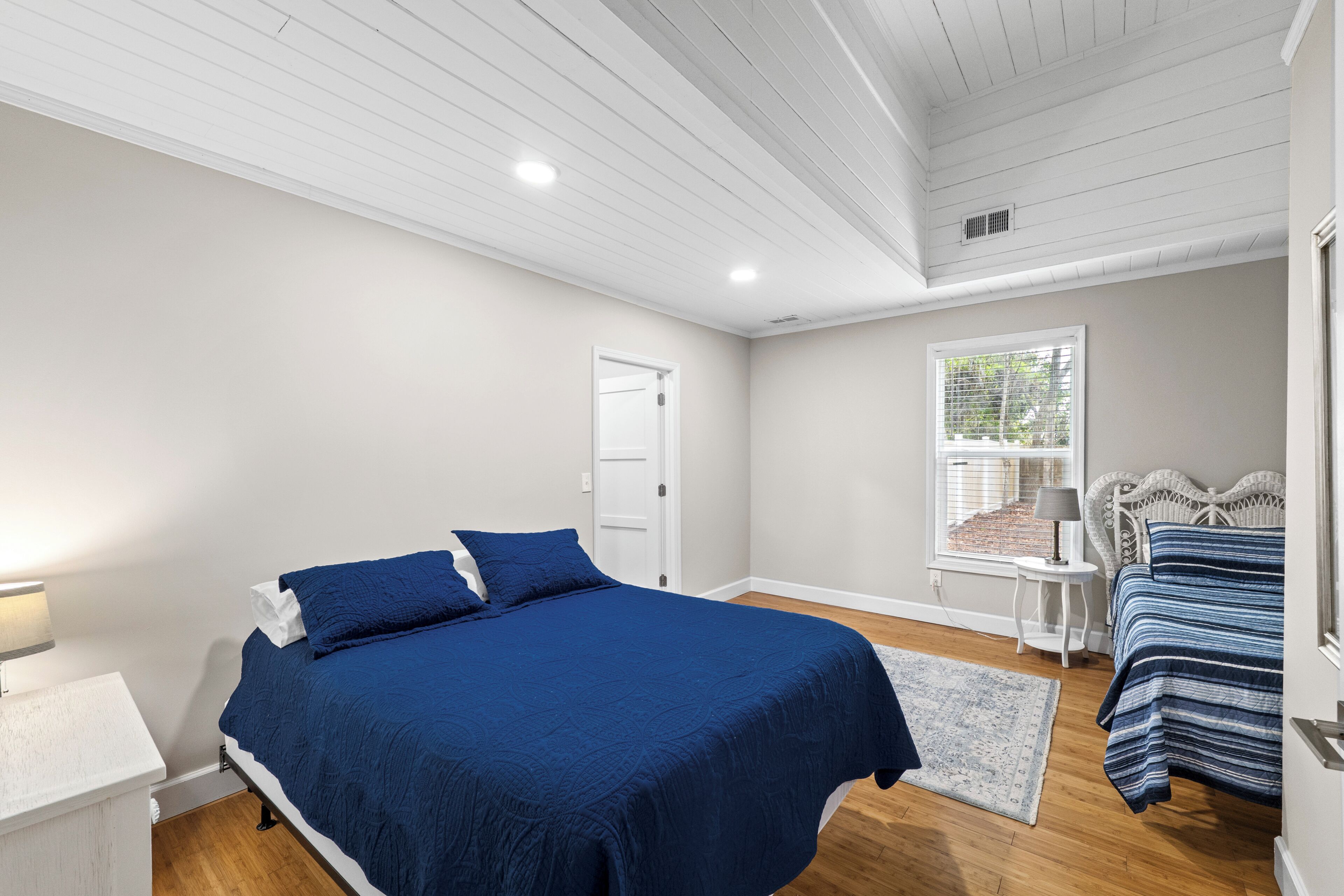 A bedroom with a large bed, a bedside table, a window with blinds, and a door.