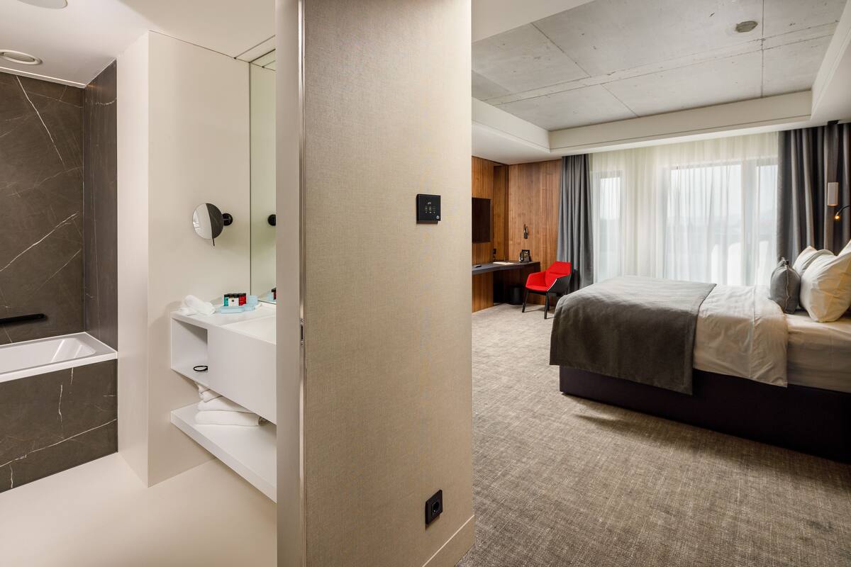A modern hotel room with a bathroom, a bed, a desk, and a view of the city.