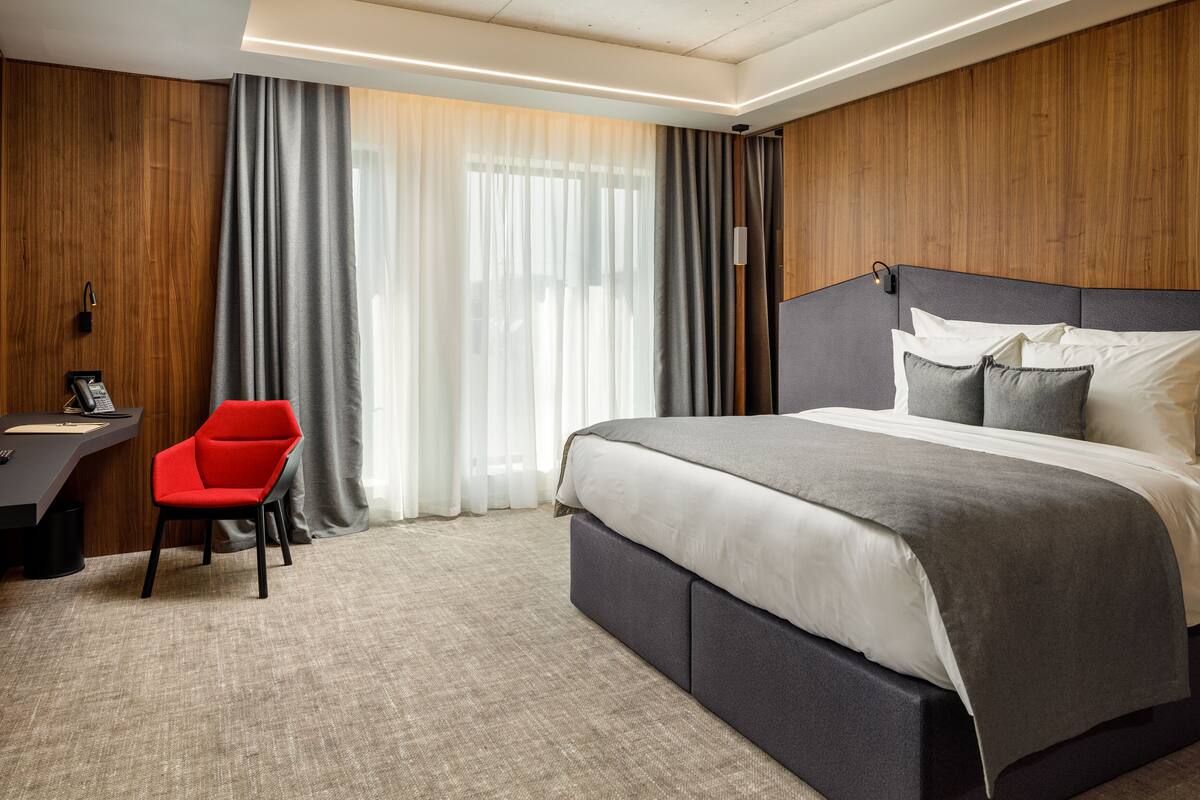 A modern hotel room with a large bed, a desk with a chair, and a view of the outdoors through sheer curtains.
