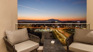 A balcony with two chairs and a table, overlooking a cityscape at dusk with visible traffic and distant mountains. - Qosmo Brasov Hotel (Brasov)