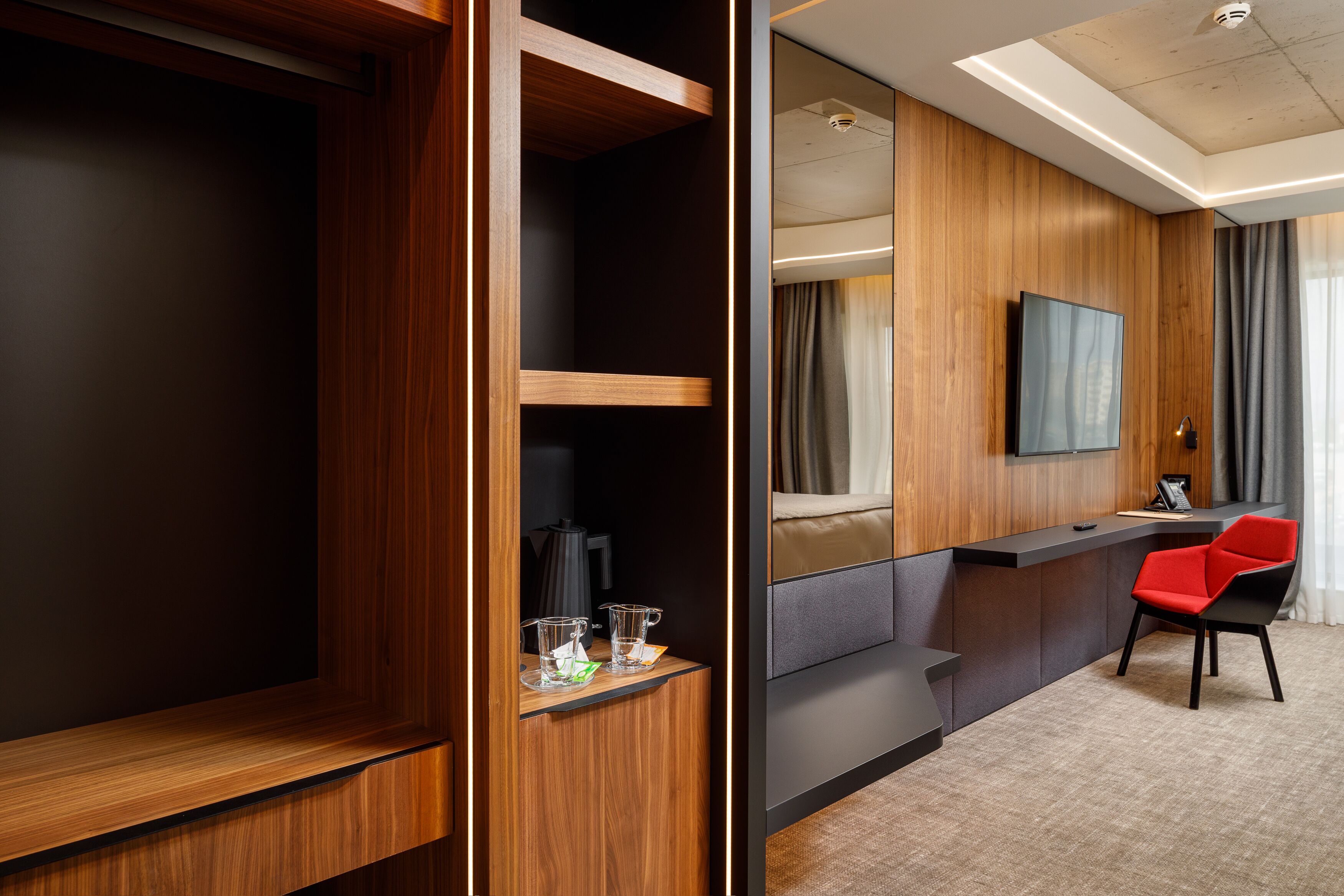 A hotel room with a wooden wardrobe, a desk with a chair, a flat-screen TV, and a large window with curtains.