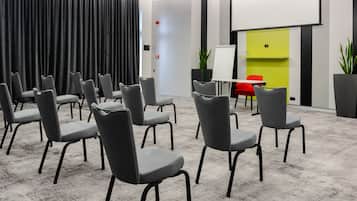 A conference room with grey chairs arranged in rows, a whiteboard, and a red table with chairs.